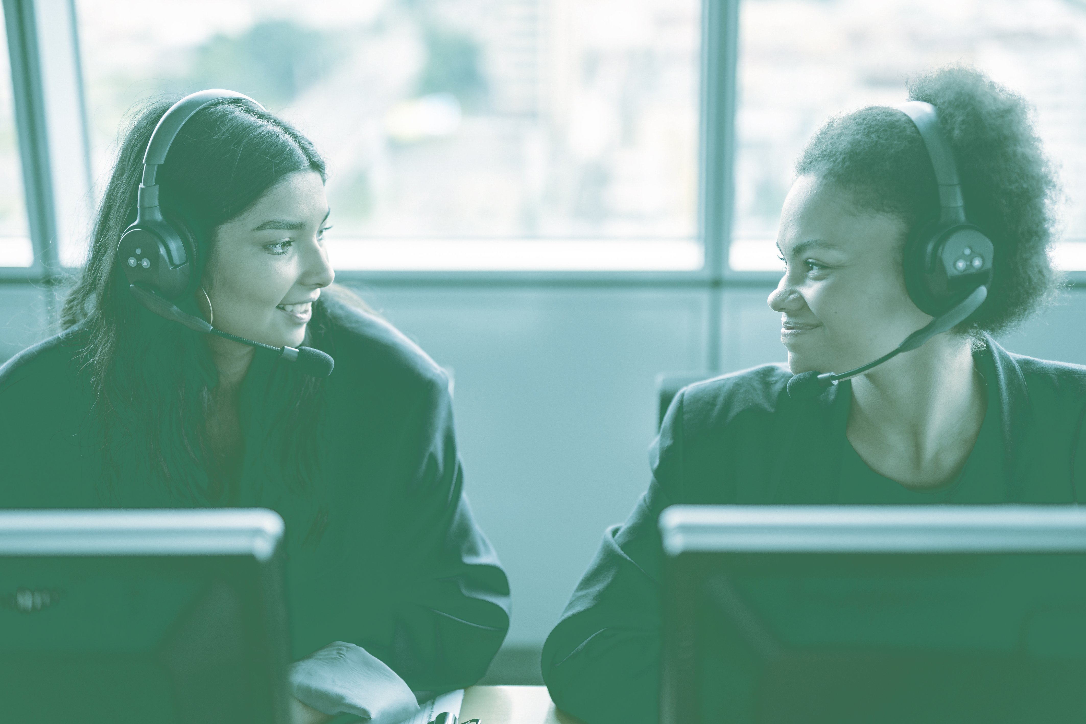 Two girls with a headset smiling each other while working on the office